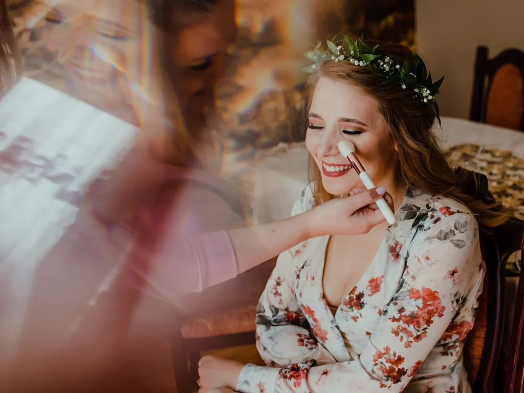 Brud under makeup-forberedelser på bryllupsdagen, smilende og omgivet af bløde farver, fotograferet af bryllupsfotograf på Sjælland nær København.