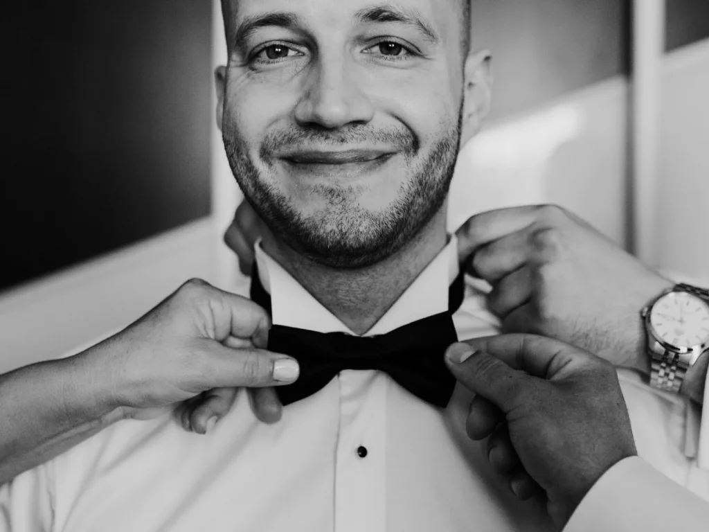 Gommen smiler, mens hans butterfly justeres af venner og familie, fotograferet i sort-hvid af bryllupsfotograf på Sjælland nær København.