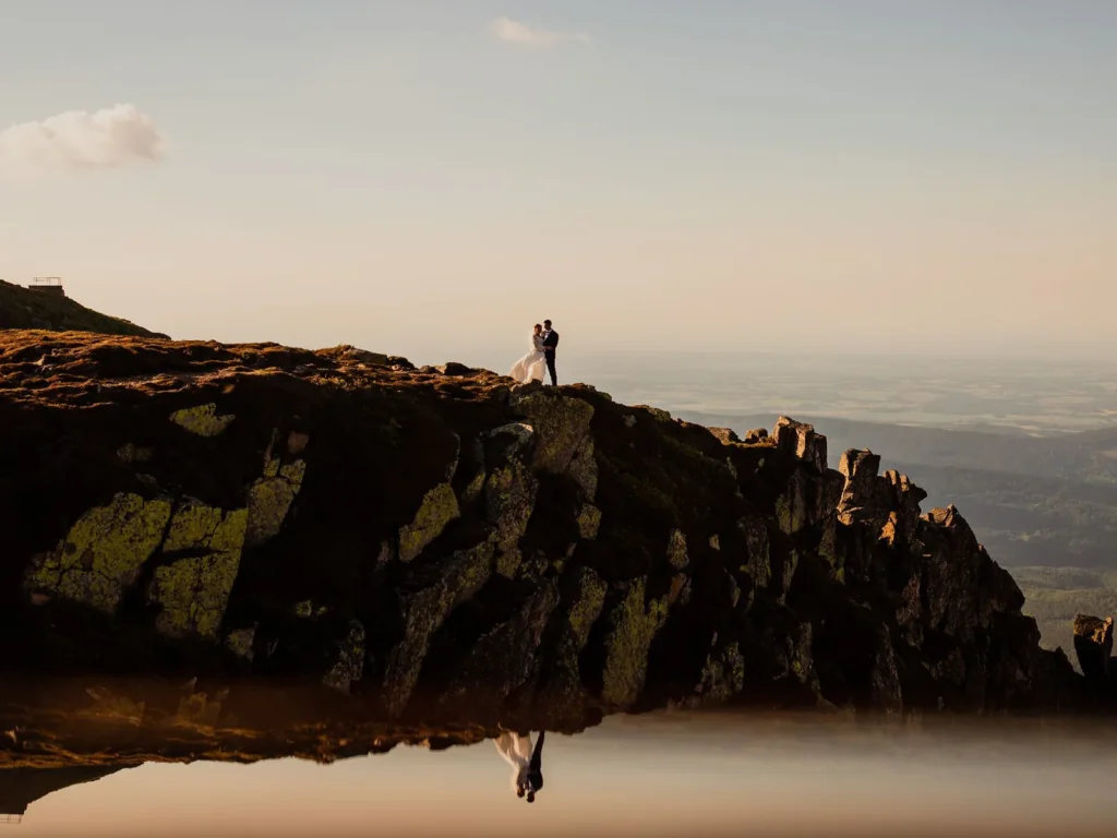 Brudepar står på en klippe med panoramaudsigt over landskabet, deres refleksion spejlet i en sø, fotograferet af bryllupsfotograf på Sjælland nær København.