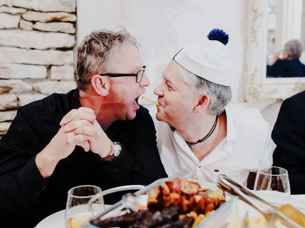 To mænd griner og har det sjovt ved bryllupsfesten, omgivet af mad og festlig stemning, fotograferet af bryllupsfotograf på Sjælland nær København.