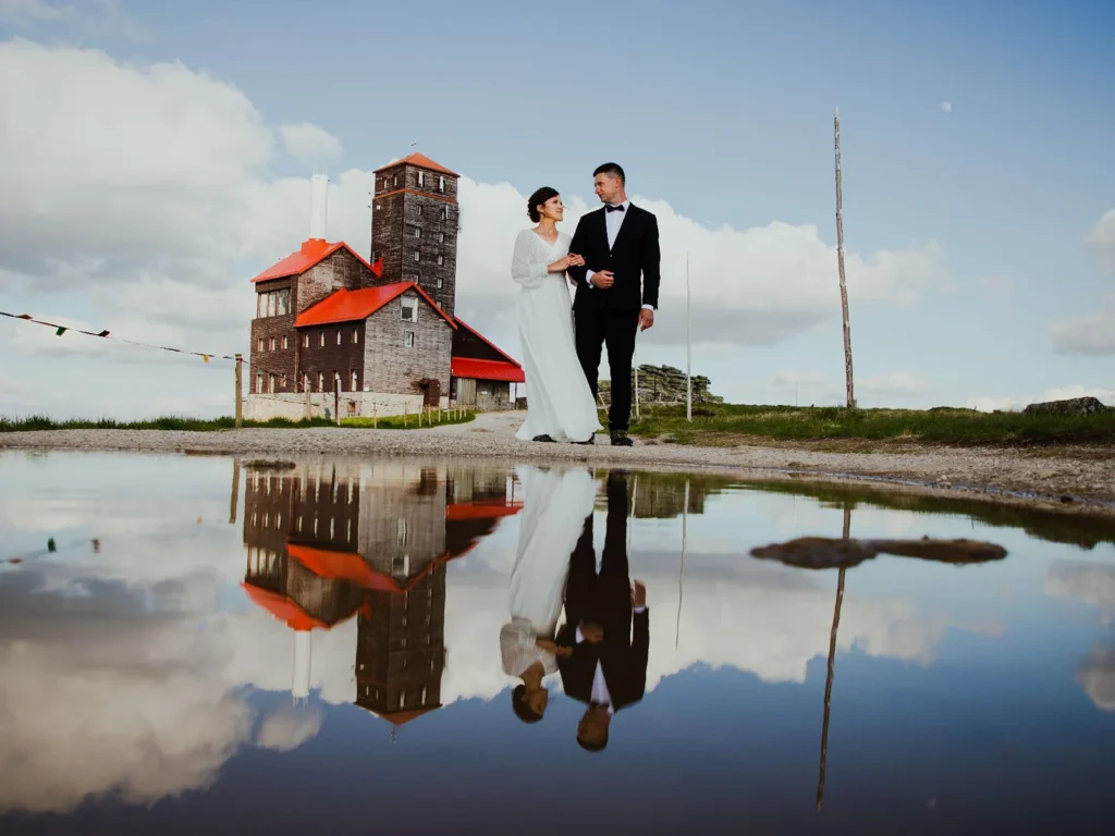 Brudepar ved en historisk bygning med rødt tag, spejlet i en vandpyt, fotograferet af bryllupsfotograf på Sjælland nær København.
