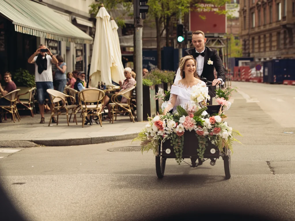 Brudepar cykler gennem byens gader i en blomsterudsmykket ladcykel, omgivet af smilende tilskuere.