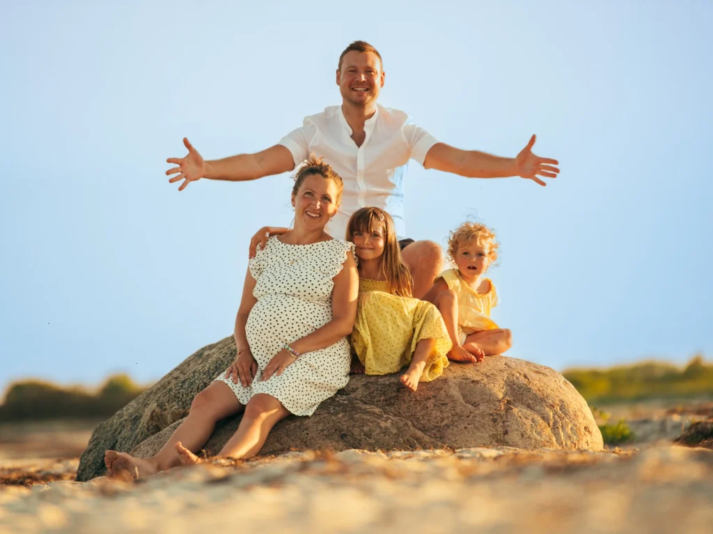 Familie sidder på en stor sten i solen med faren, der strækker armene ud – smukke og naturlige familiebilleder fra en familiefotograf i København og på Sjælland.