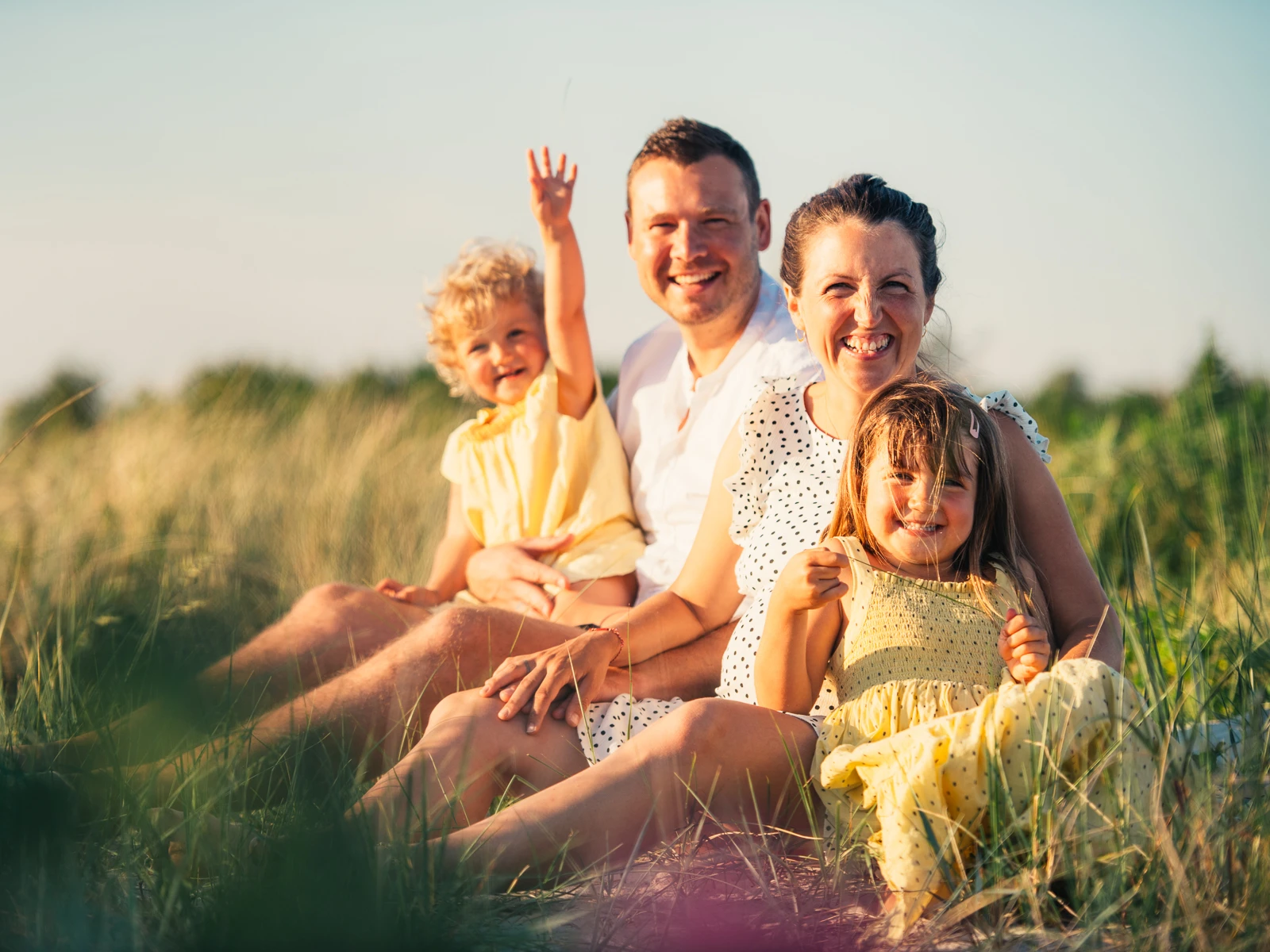 Familiefoto med glæde og nærvær – Familiefotografering på Sjælland Smilende familie i solen omgivet af græs og natur – livsglade familiebilleder fra en familiefotograf i København og på Sjælland.
