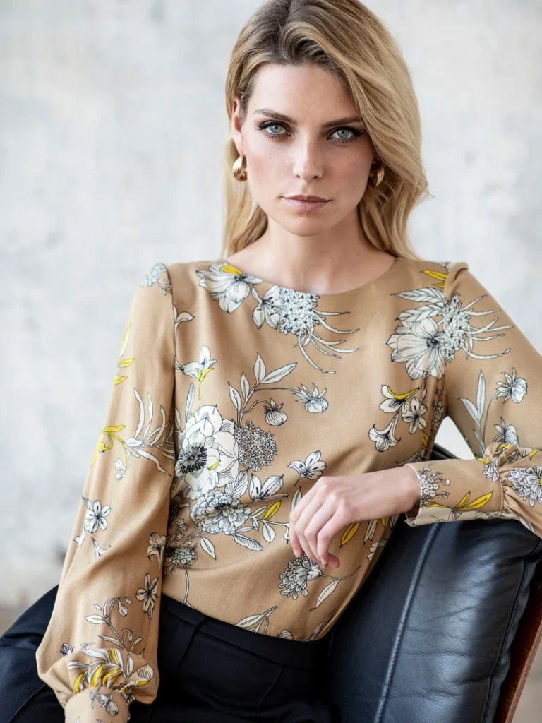 Kvinde i en beige bluse med blomsterprint og sorte bukser sidder på en stol foran en betonvæg.