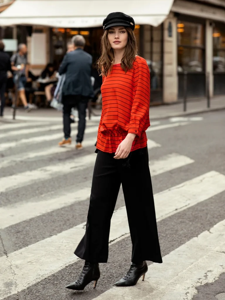 Kvinde i rødstribet bluse og sort kasket krydser gaden i et urbant miljø med cafébaggrund.