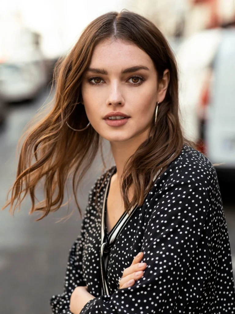 Portræt af en kvinde med brunt hår iført en sort bluse med hvide prikker, fotograferet i bymiljø.