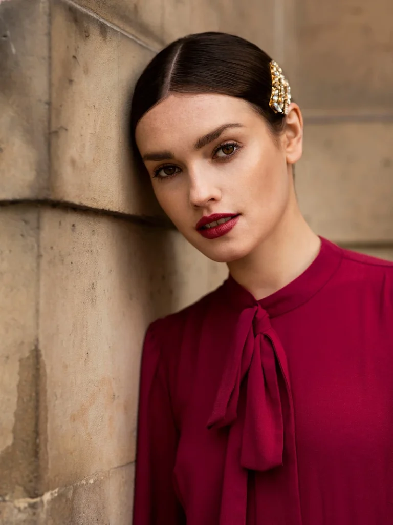 Portræt af en kvinde i en dyb rød bluse med en elegant hårspænde, lænet op ad en stenvæg.