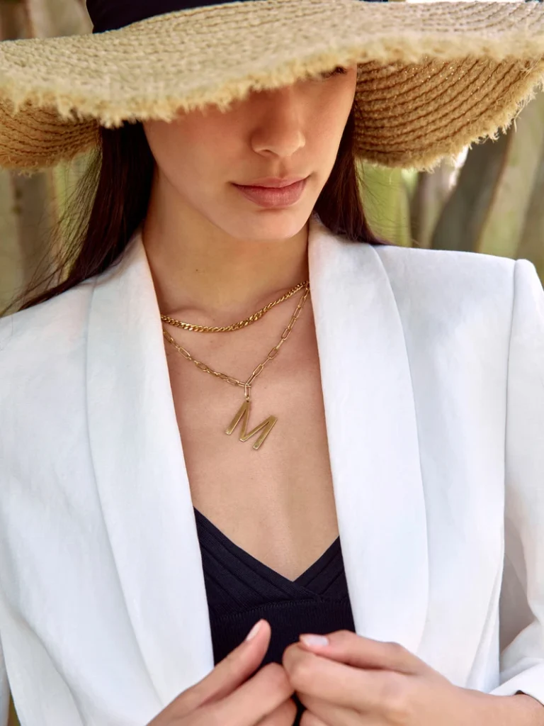 Detaljebillede af en model i hvid blazer med stråhat og gyldne halskæder.