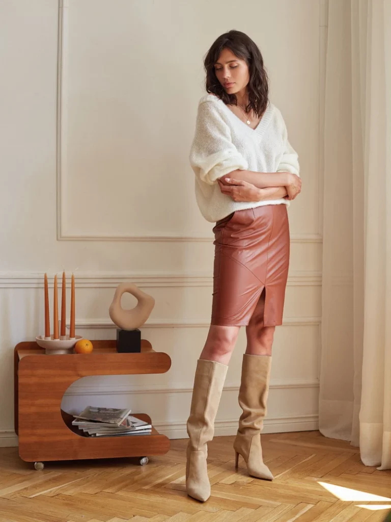 Modefotografi af en kvinde i hvid strikbluse, brun lædernederdel og beige støvler, stående i et minimalistisk indrettet rum.
