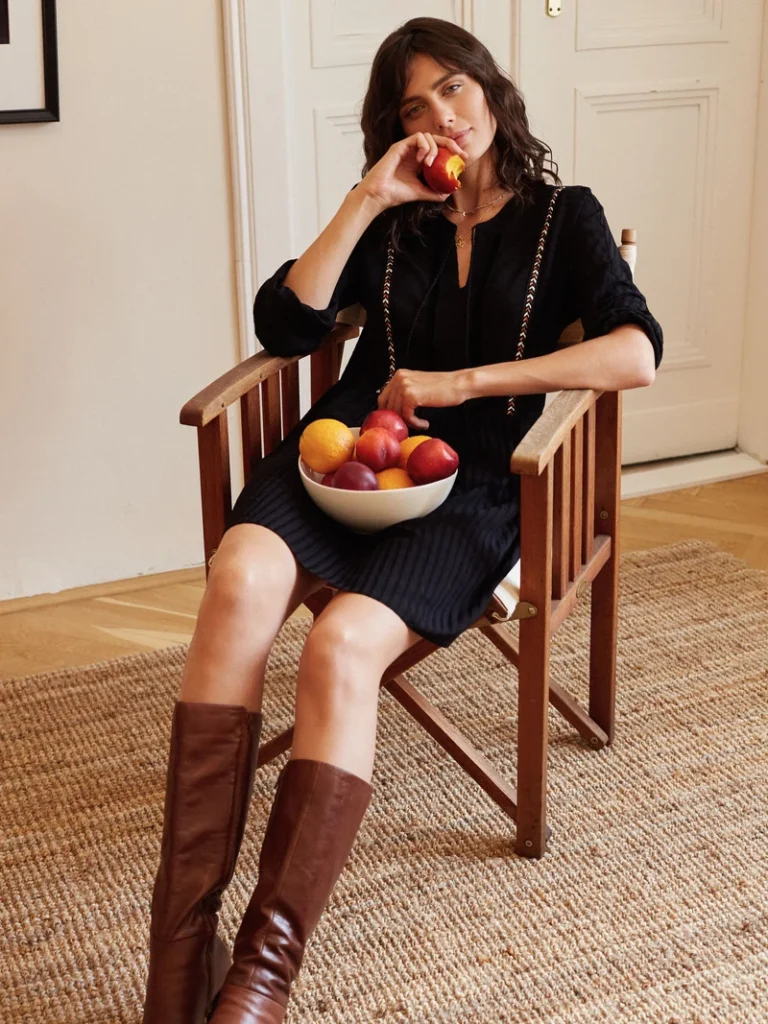 Modefotografi af en afslappet kvinde i sort kjole og lange brune støvler, siddende i en stol med en skål frugt i skødet.