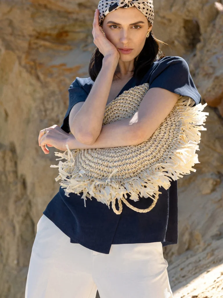 Model i mørkeblå bluse og hvidt tøj med en flettet stråtaske og prikket tørklæde foran en stenbaggrund.