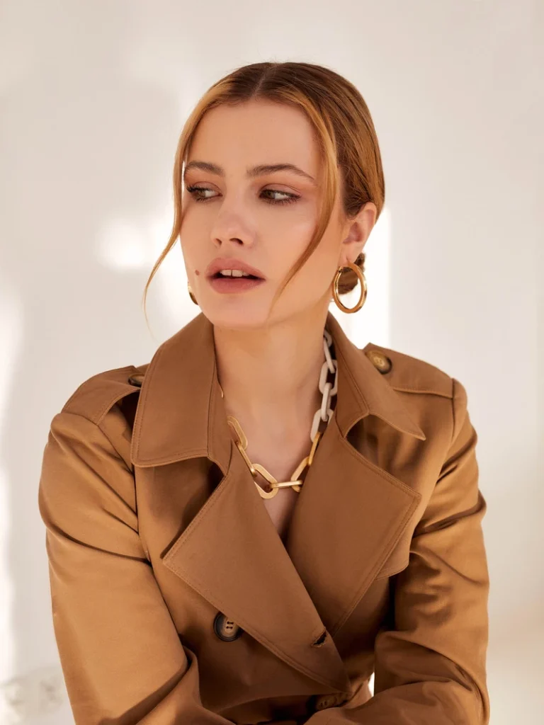 Portræt af en kvinde i camel trenchcoat med gyldne accessories, badet i varmt naturligt lys.